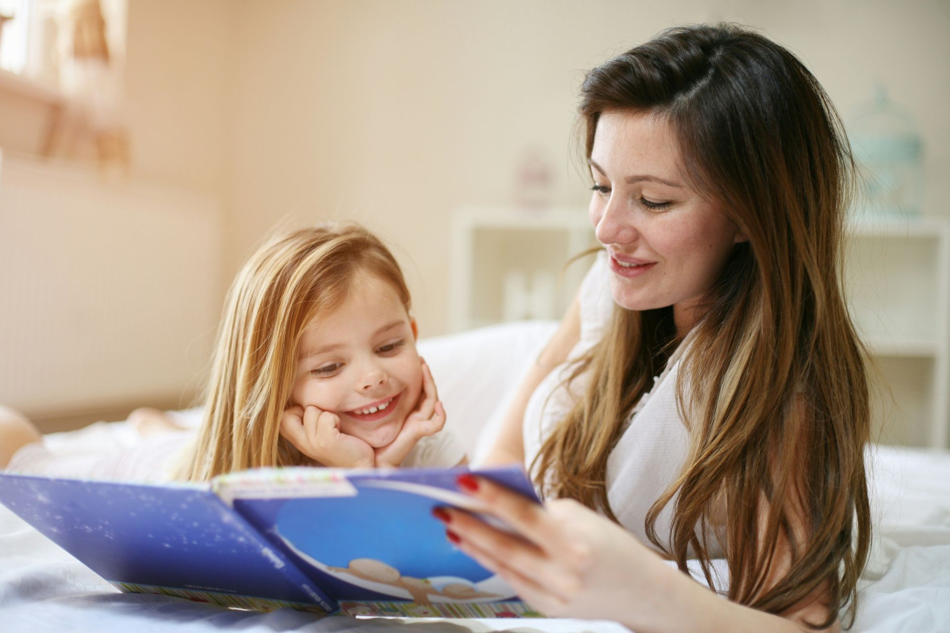 Mother and elementary-aged daughter sitting together on a couch and smiling as they share a book.