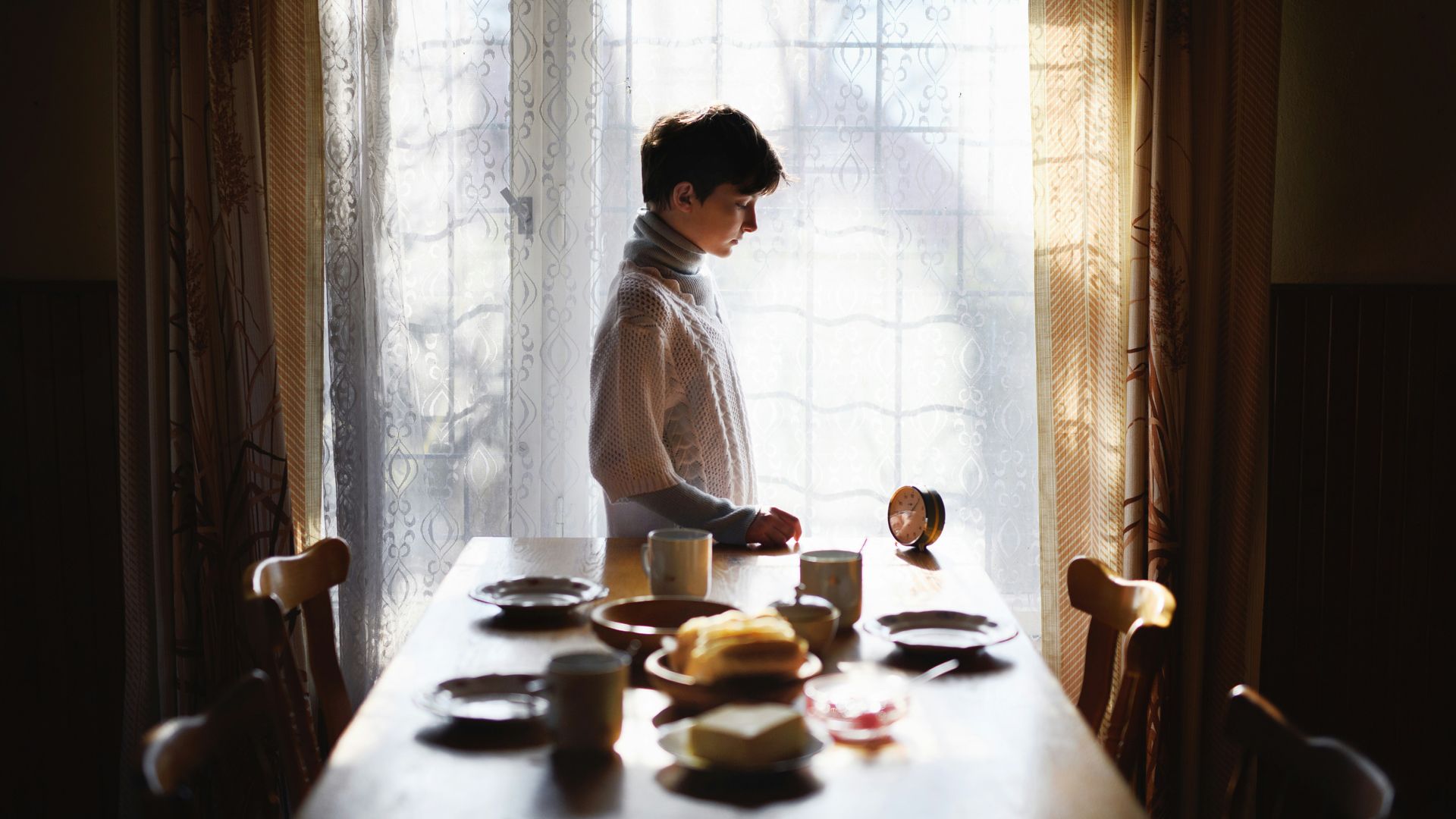 Woman standing alone beside a dining table set for several people, looking down as sunlight shines through a large window.