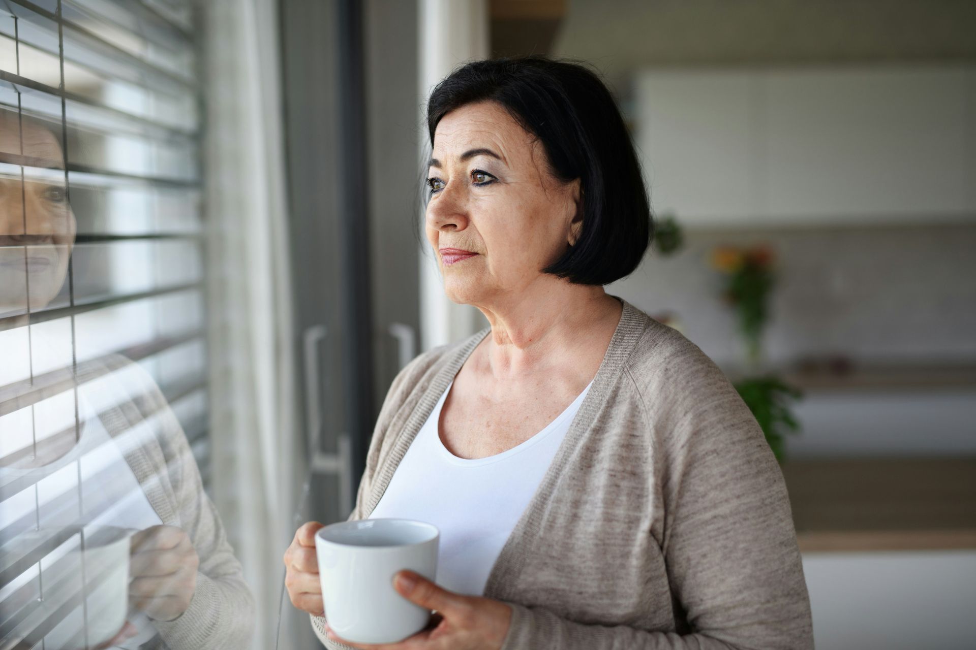 Middle-aged woman, holding a coffee cup while looking out a window, reflecting on her past.