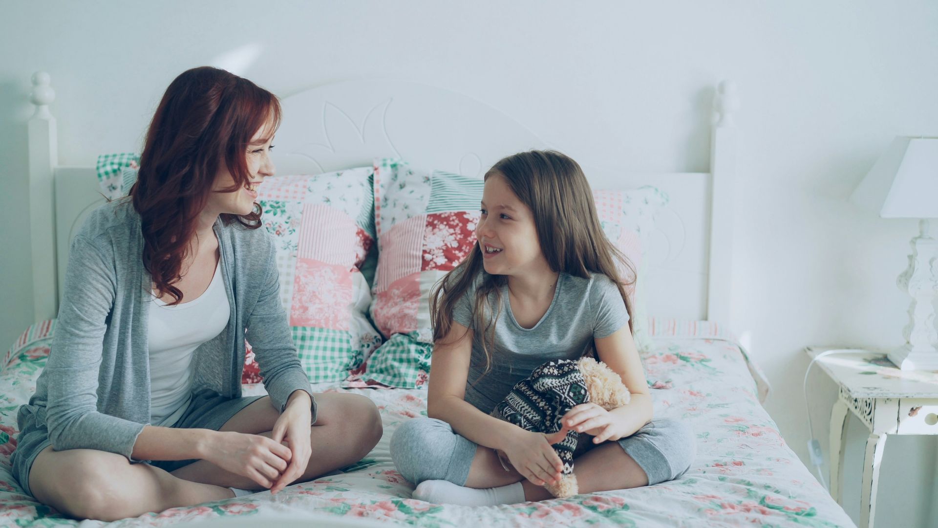 Mother and daughter are talking conversationally while sitting on the daughter's bed.