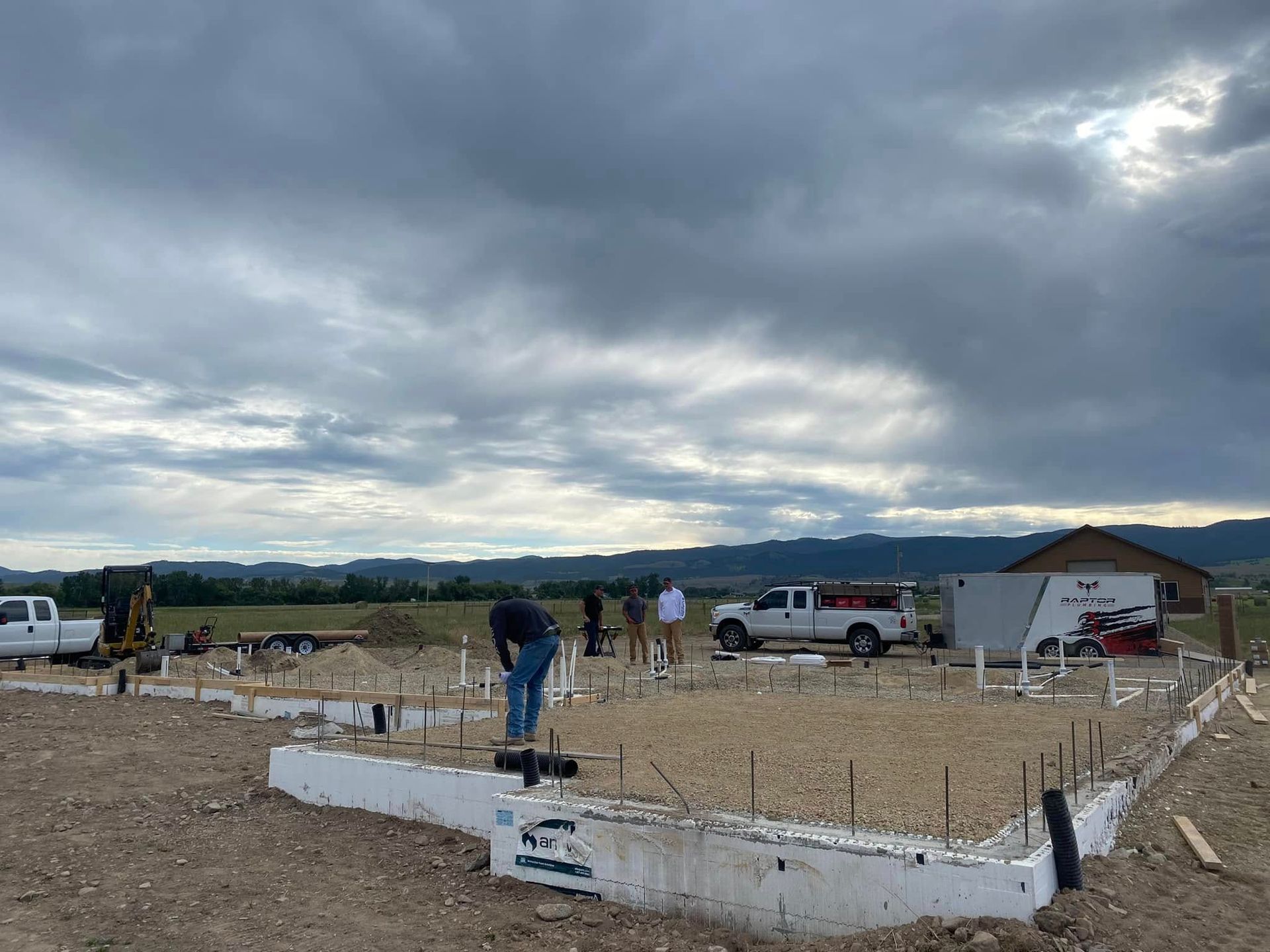 Concrete foundation under construction on a rural lot, with workers, trucks, and a cloudy sky.