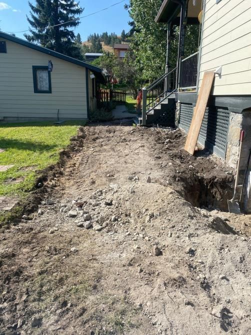 Dirt trench beside a house, with a narrow yard and a board leaning against the foundation.