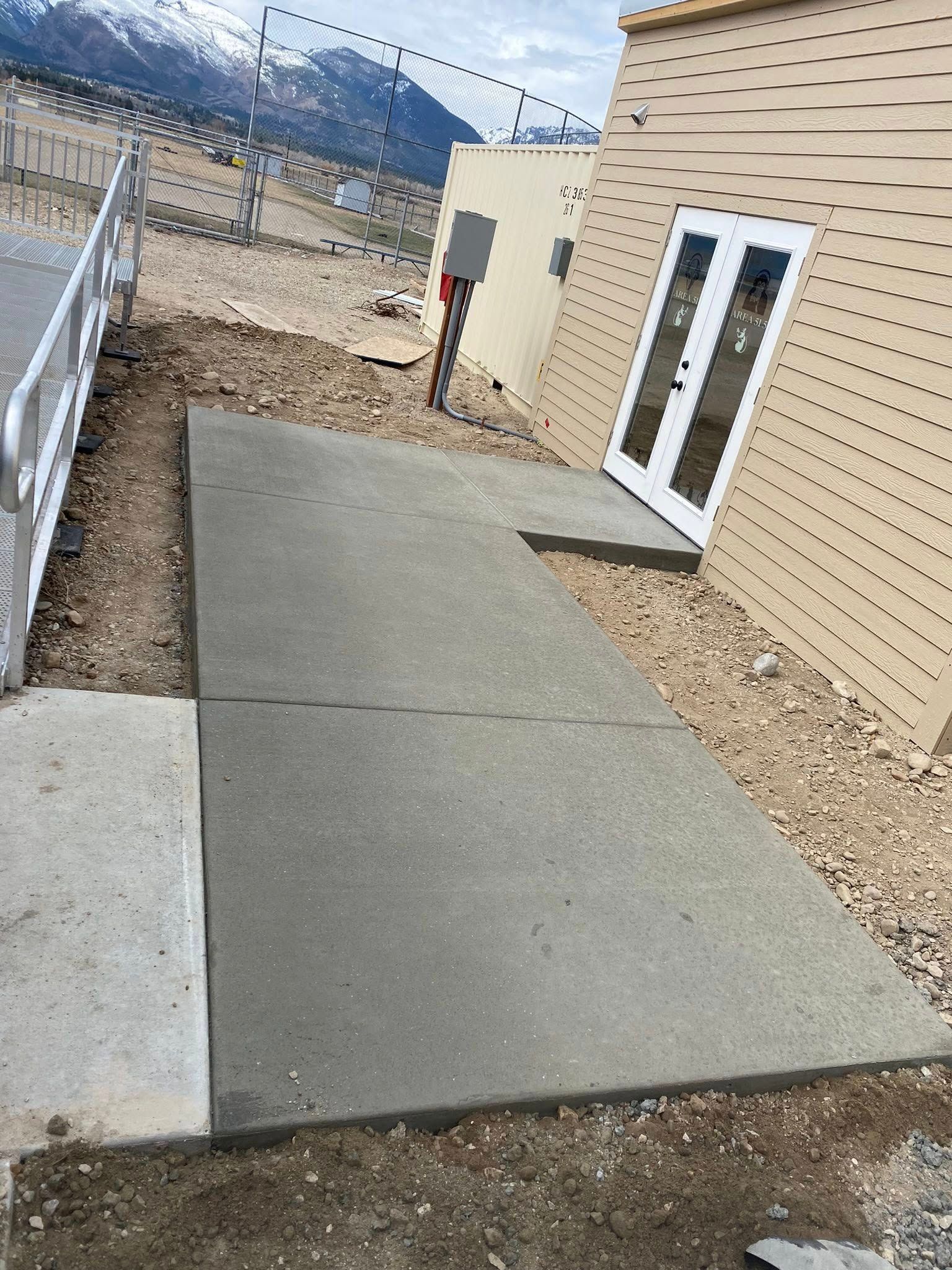 Concrete walkway beside a beige building with white double doors, bordered by gravel and a metal railing.