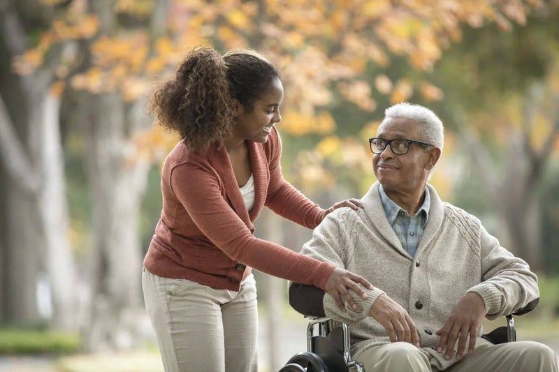 Woman in a coral sweater helps an older person in a wheelchair outdoors among autumn trees