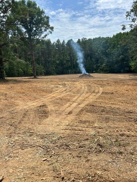 Cleared dirt field with a small smoke plume in a wooded area under a partly cloudy sky