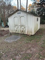Weathered white wooden shed with double doors in a wooded yard.