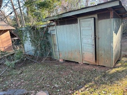 Weathered gray shed with a white door beside an overgrown yard and trees