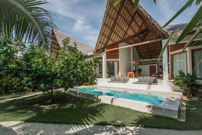 A thatched-roof villa with a private swimming pool, green lawn, and tropical plants under a blue sky.