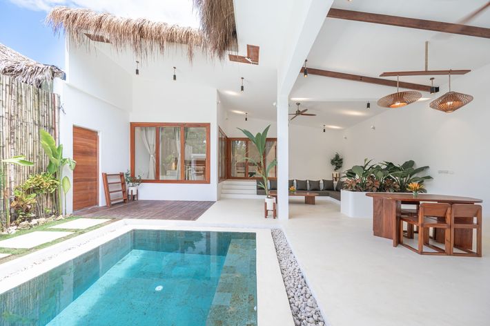 A modern, open-air villa with a pool, wooden dining furniture, white walls, and a thatched roof accent.