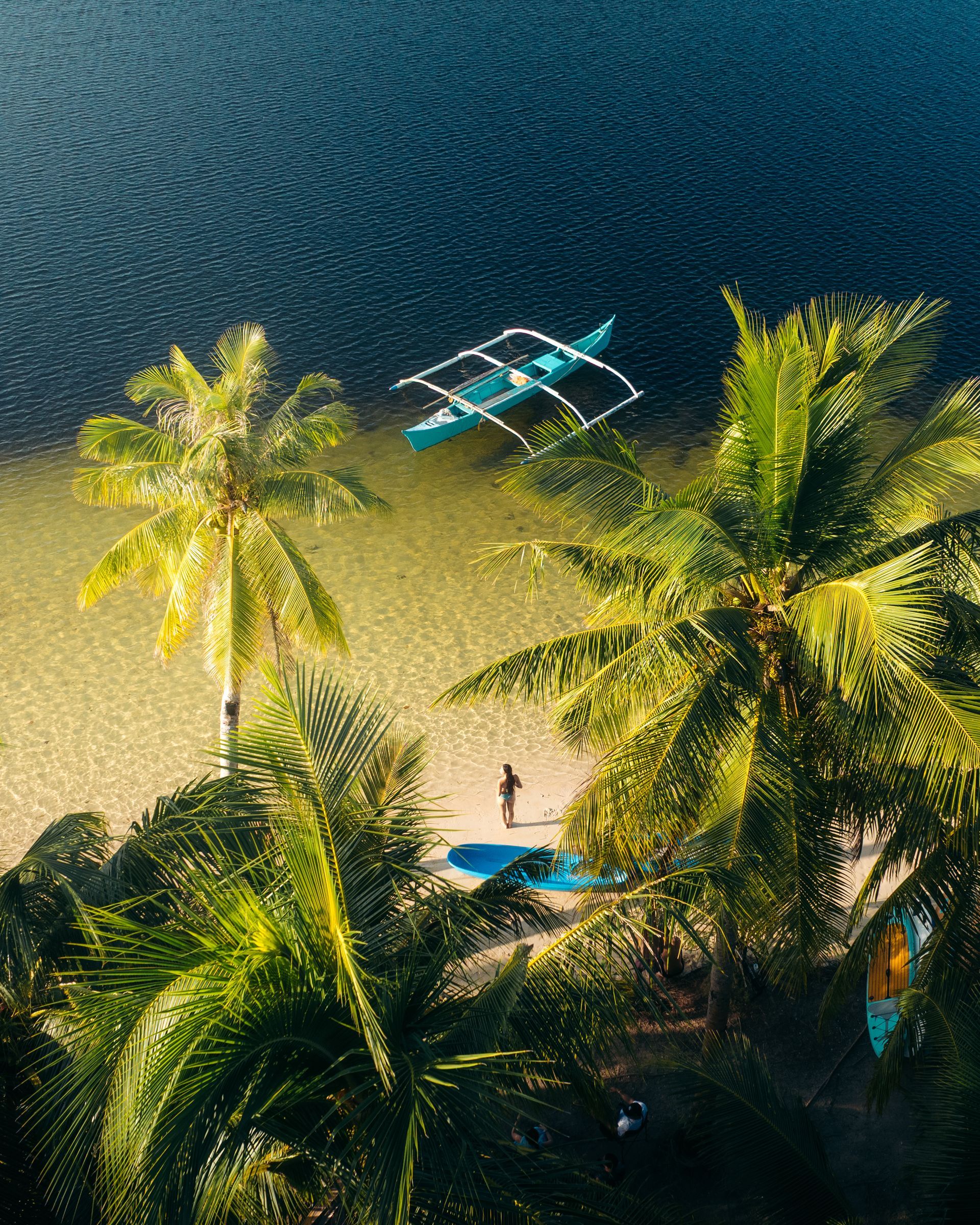 A high-angle view of a sandy beach with palm trees, a small traditional boat in clear water, and a person standing below.