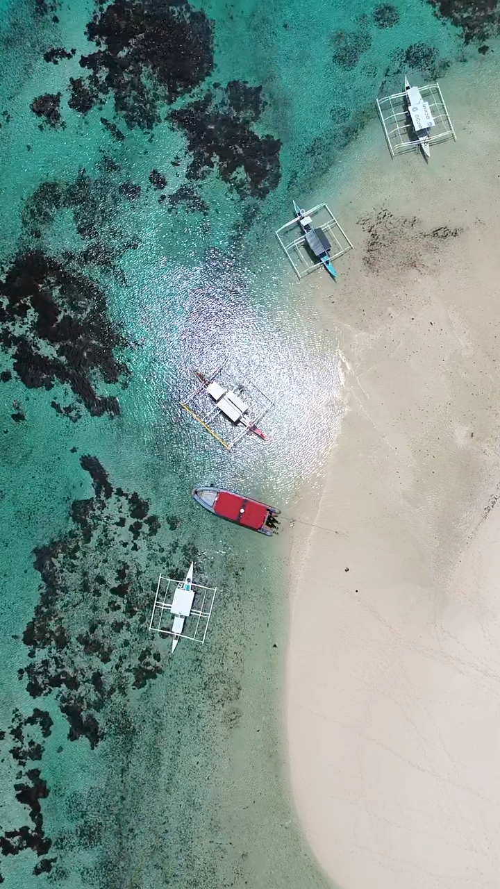 Aerial view of five boats anchored in clear turquoise water beside a white sandy beach with dark rock formations nearby.