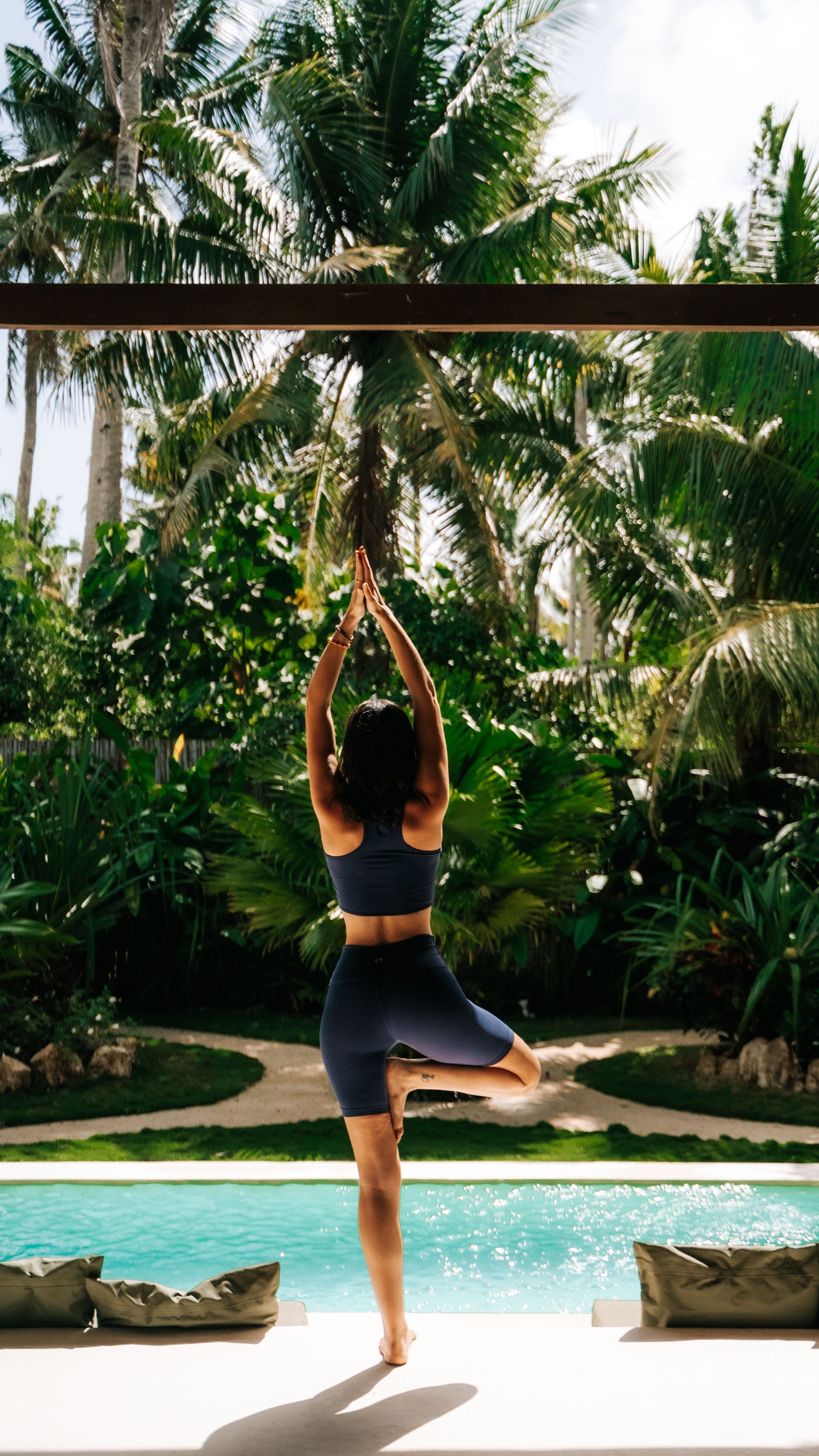A person in dark activewear performs a yoga tree pose by a swimming pool surrounded by lush tropical greenery.