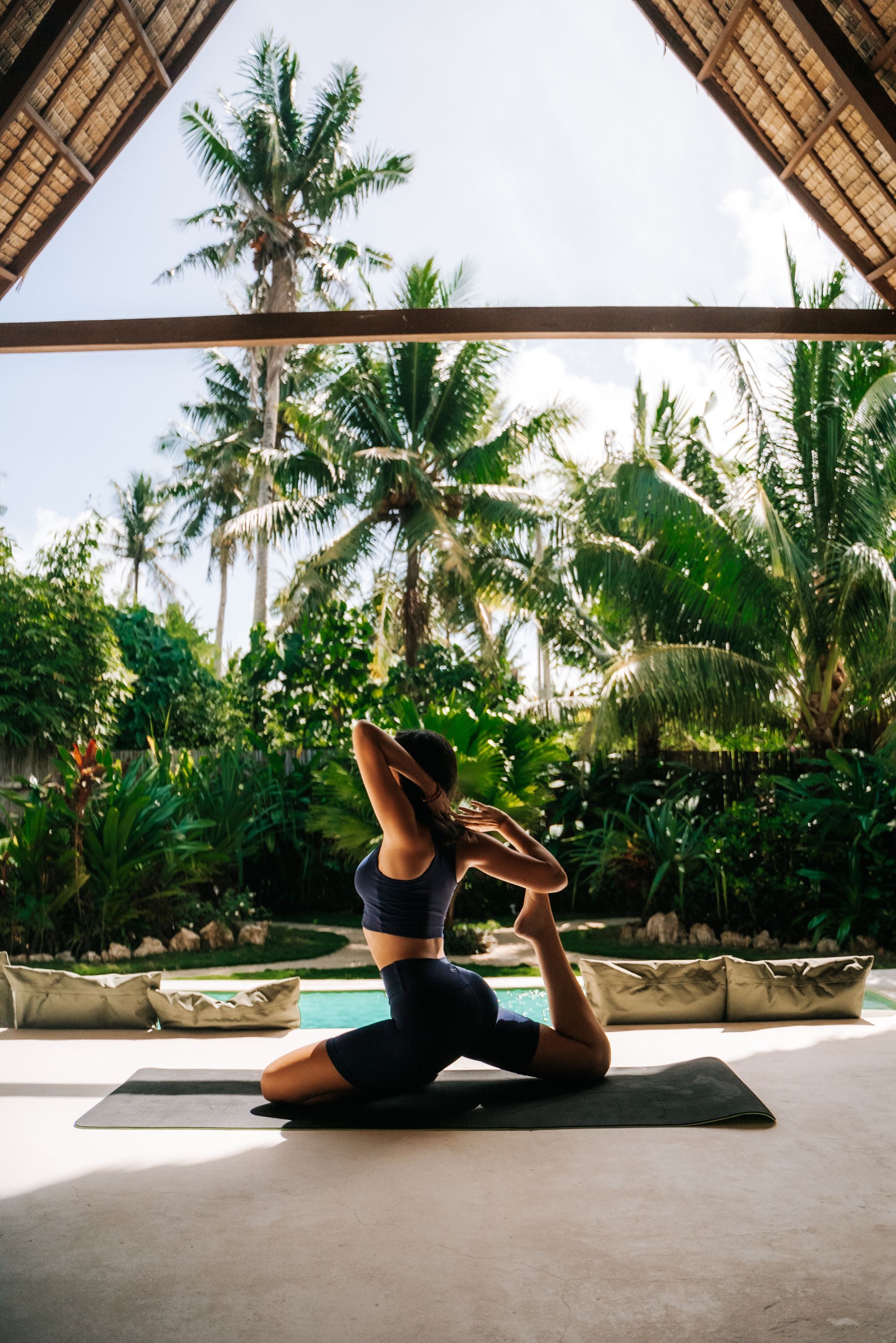 A person in dark activewear performs a mermaid yoga pose on a mat in an open-air tropical pavilion overlooking a pool.