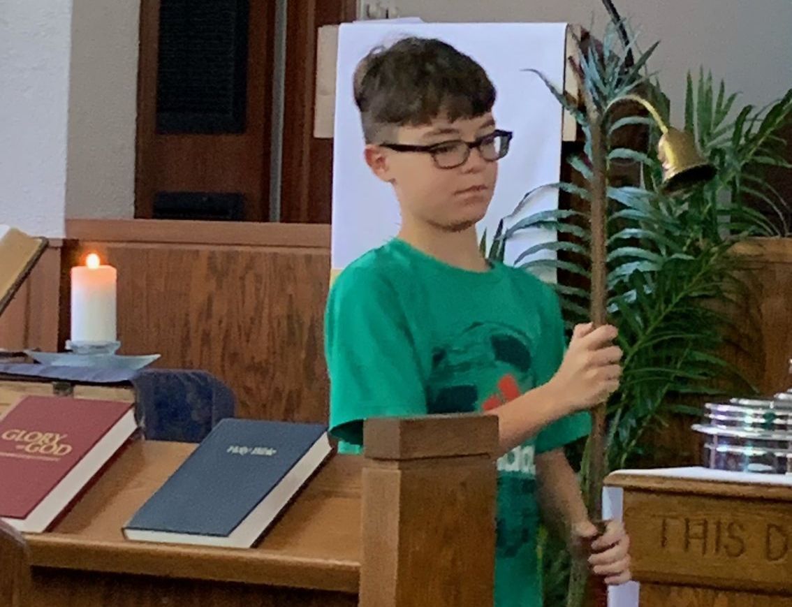 Boy in green shirt holding a tall candle beside a church altar with books and a lit candle.