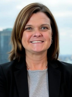 A smiling professional with shoulder-length brown hair, wearing a black blazer and a grey shirt