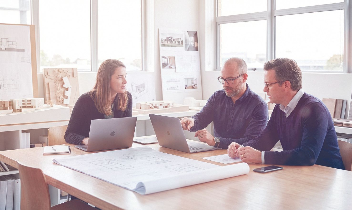 Drei Kollegen arbeiten in einem hellen Büro gemeinsam an einem Tisch mit Laptops und Architekturplänen. Bild ist mit KI erstellt