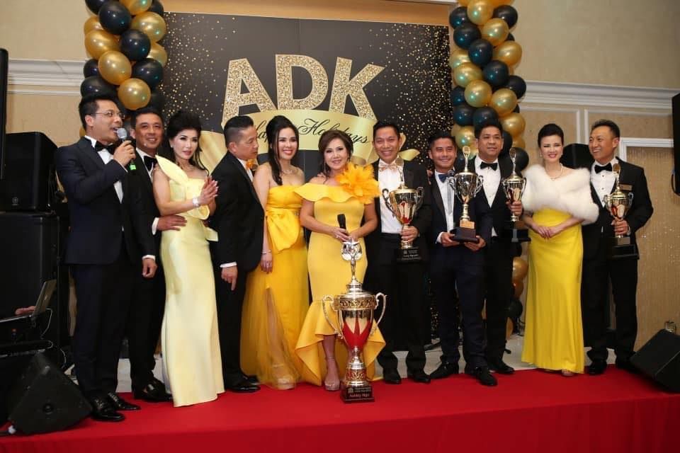 A group of people in formal attire stand on a stage before an ADK sign, some holding trophies.