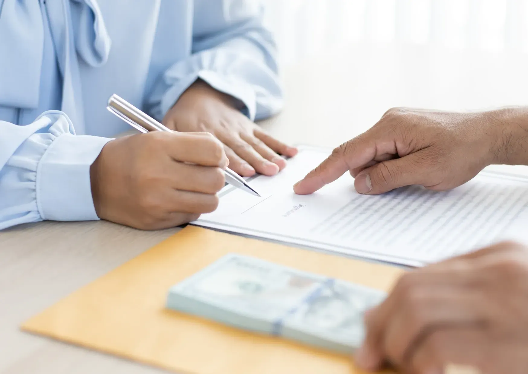 Hands signing a document at a table with a pen and cash in the foreground