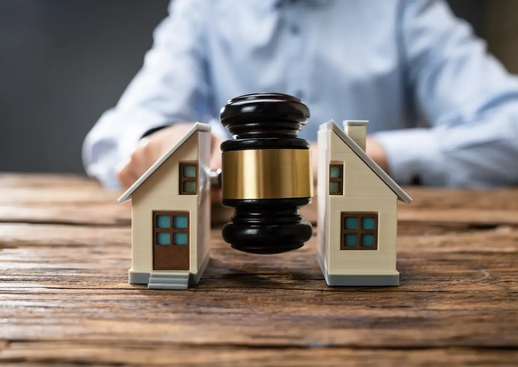 Judge’s gavel between two small houses on a wooden table, with a blurred person in the background