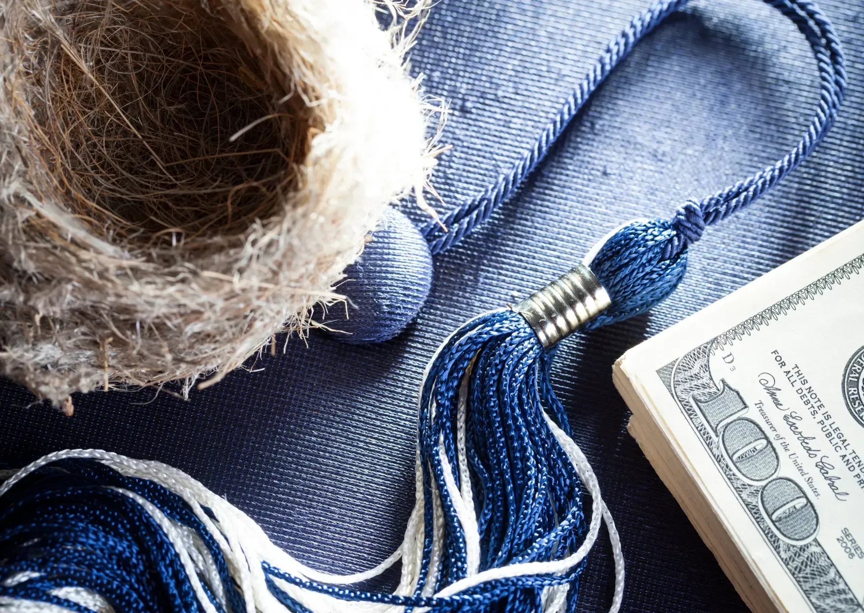 Blue graduation cap tassel beside a frayed rope and a stack of cash on denim fabric.