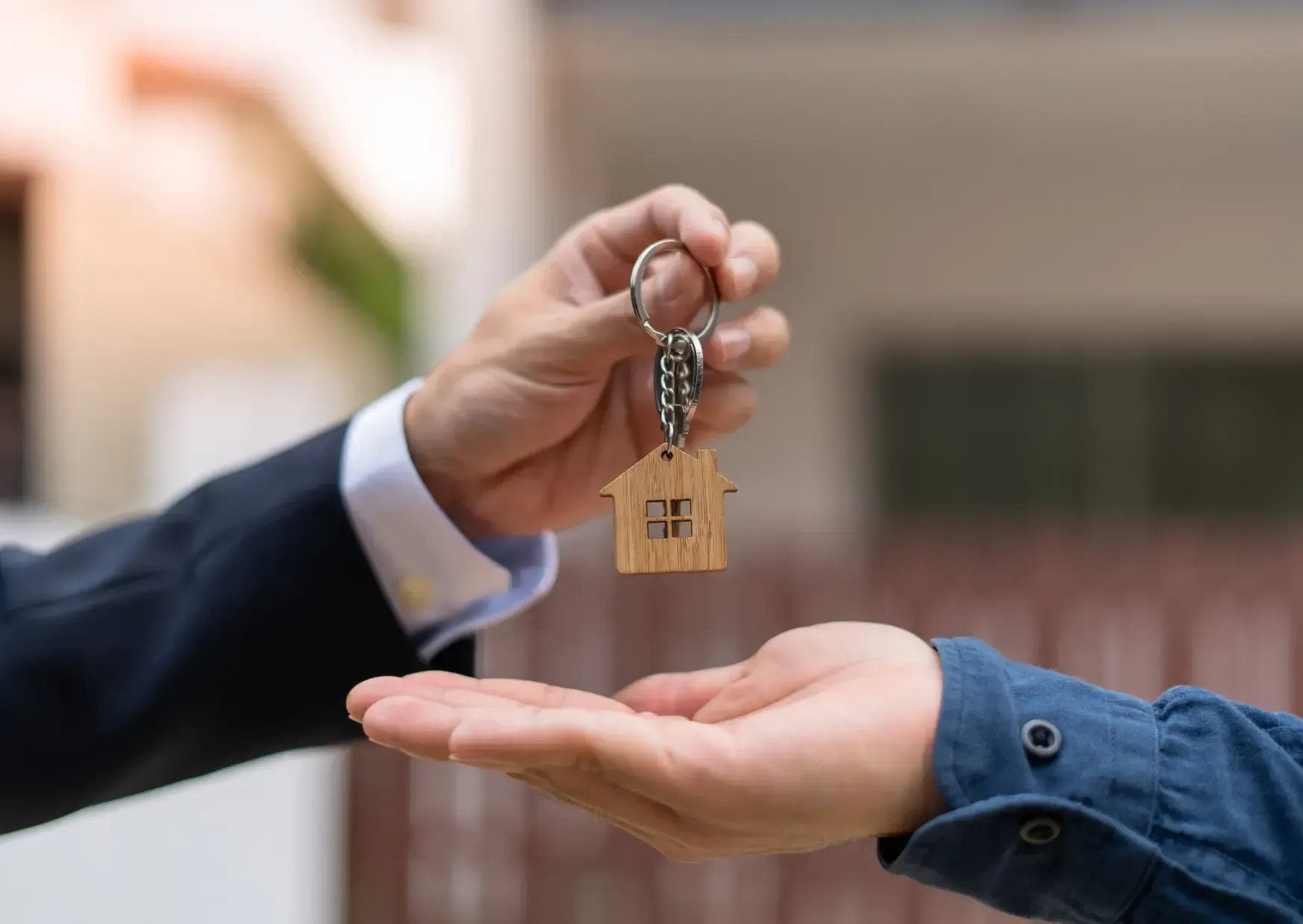Hand passing house keys to another over an outstretched hand, with a blurred home in the background
