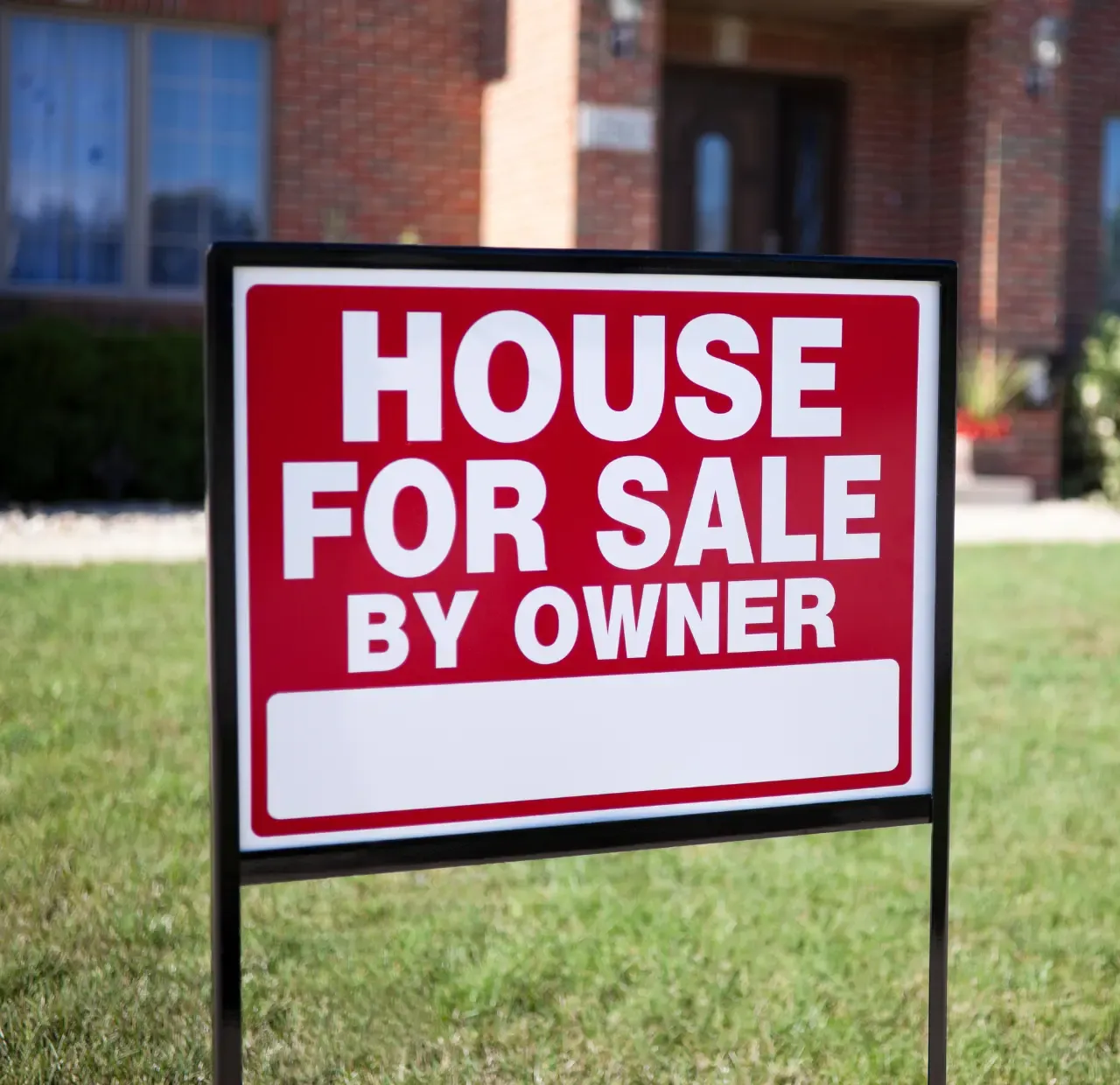 Red “House For Sale By Owner” sign on a lawn in front of a brick house.