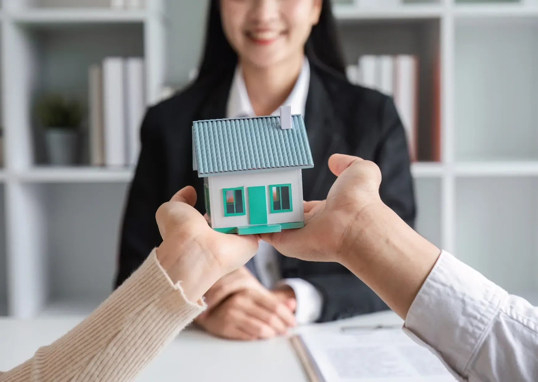 Hands holding a small teal house model in front of a smiling person in an office setting