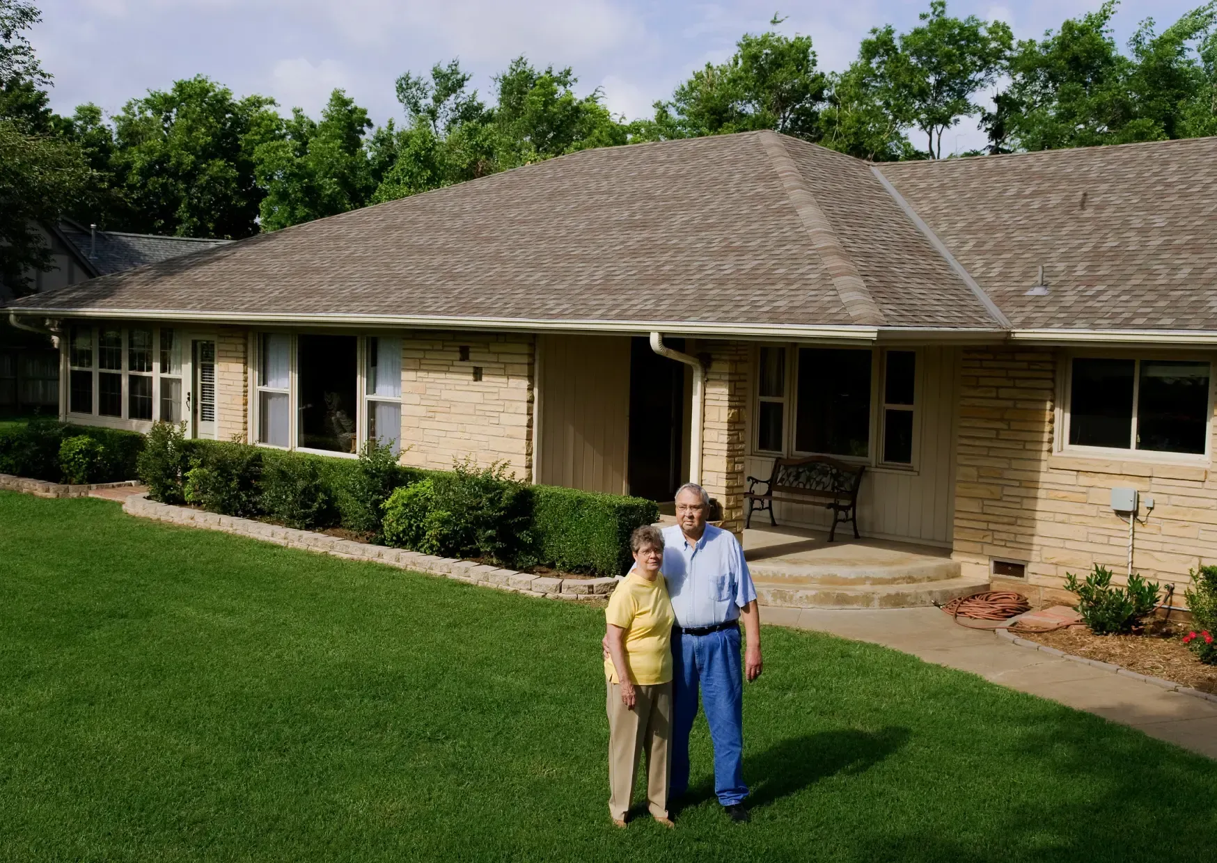 Couple standing on lawn in front of a single-story house with beige brick and a shingled roof