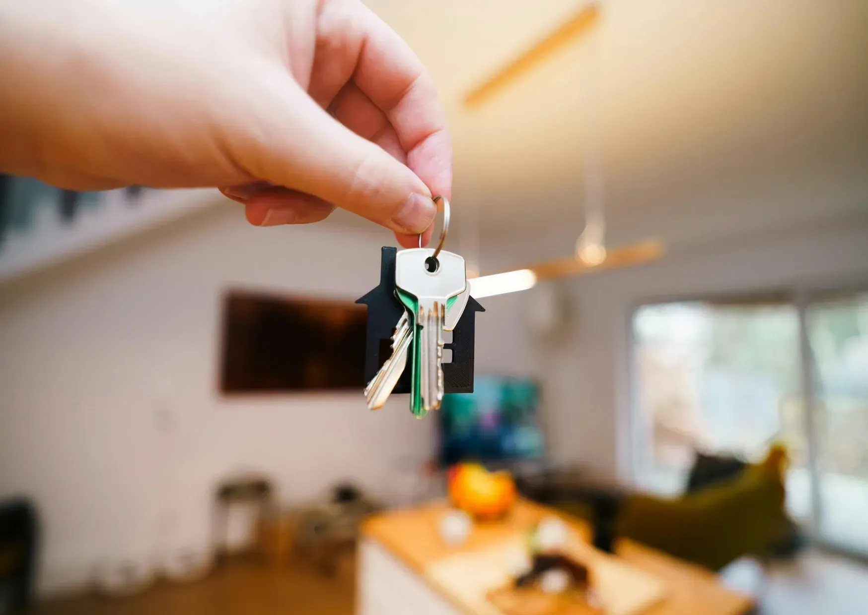 Hand holding a small house key in a bright living room with a blurred sofa and window in the background