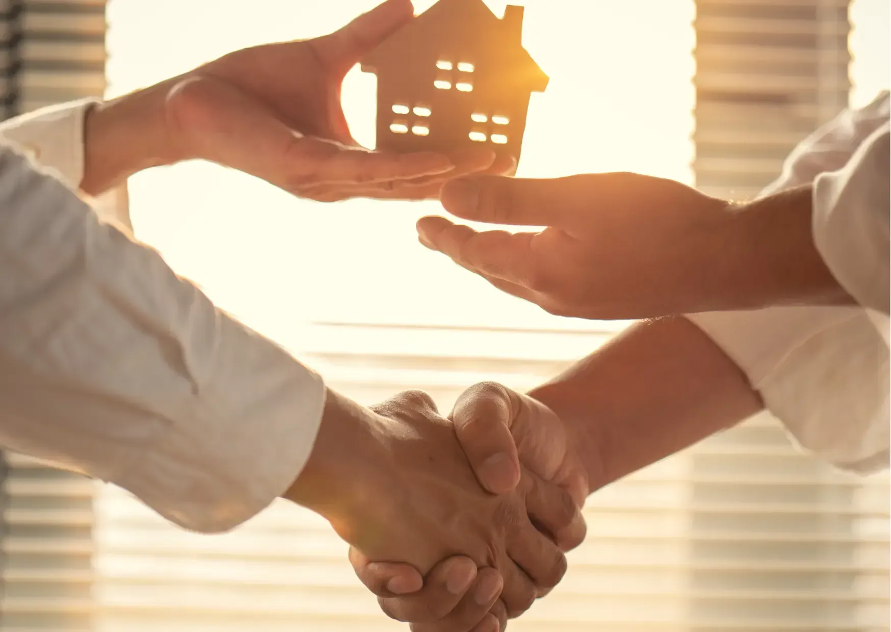 Hands shaking in front of a glowing house symbol at sunset, suggesting home sale or support