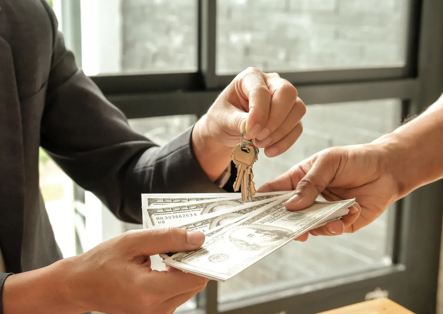 Hands exchanging keys and cash for a house sale near a window.