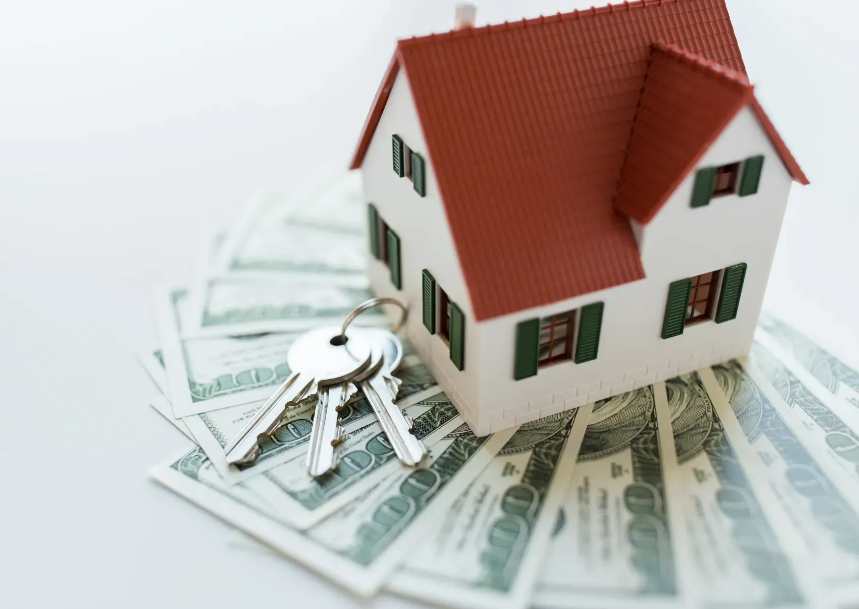 Model house with red roof sitting on scattered U.S. dollar bills and keys