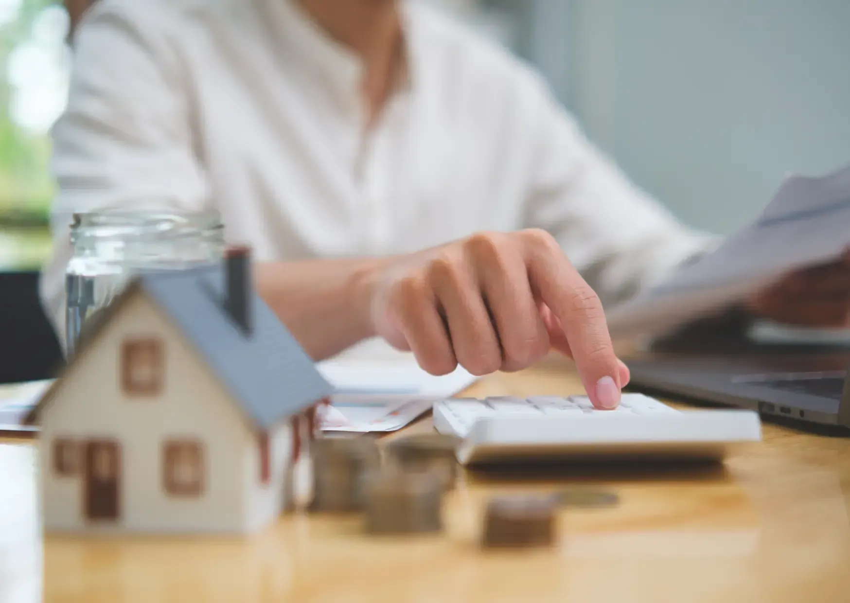 Hand pointing at documents beside a small model house and jar on a desk