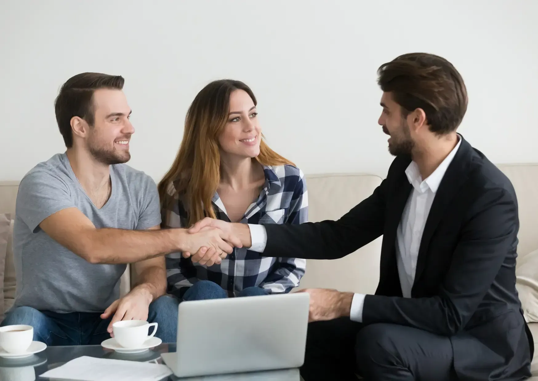 Three people in business attire shaking hands on a couch, with a laptop and coffee cups nearby.