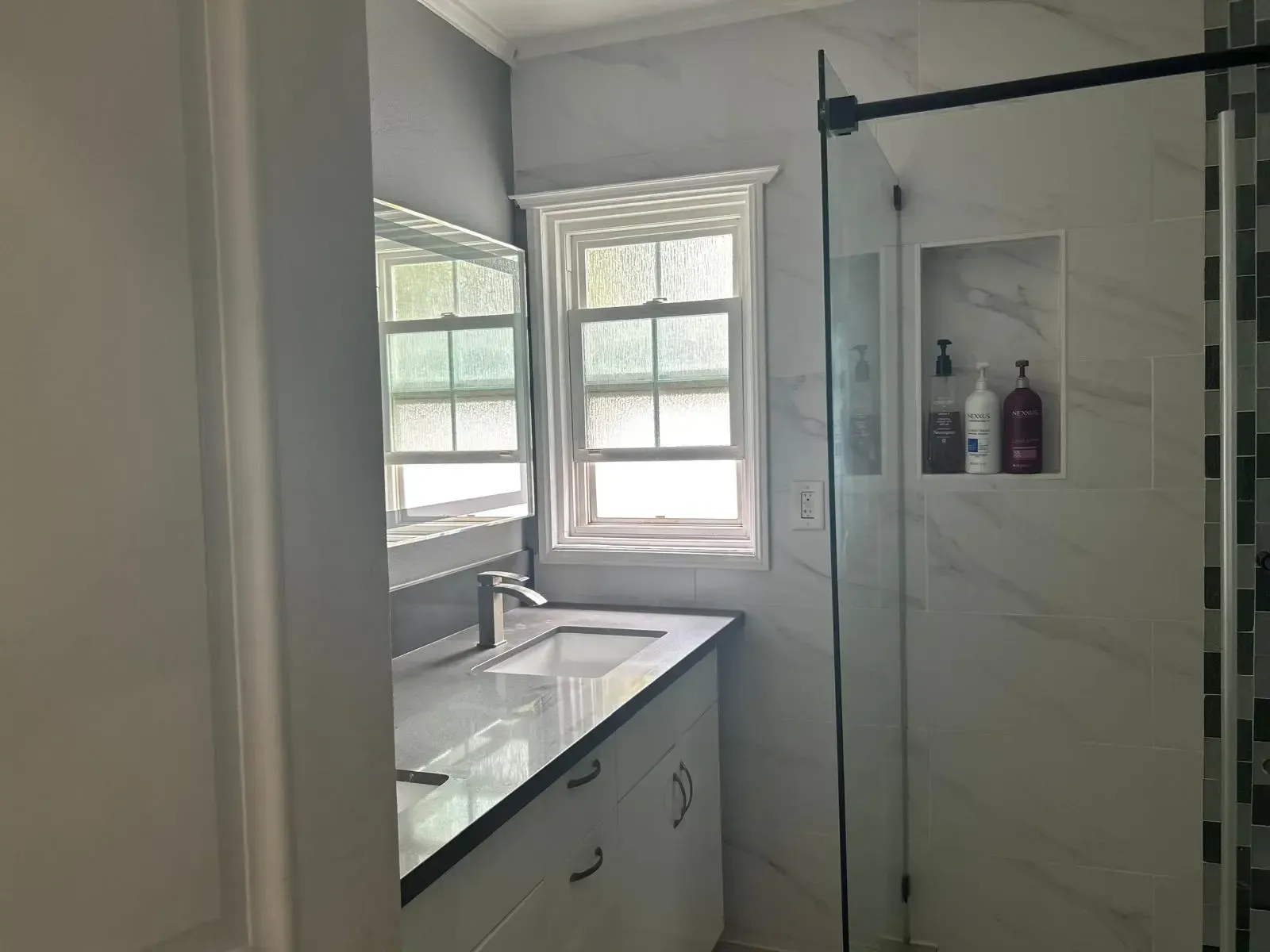 A bathroom with a sink , mirror , shower and window.