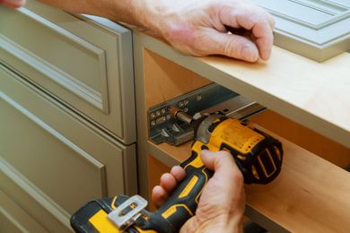 Person using a yellow drill to install a metal drawer slide inside a kitchen cabinet.
