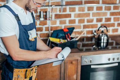 Handyman in overalls and safety glasses writing on a clipboard in a kitchen with tools.