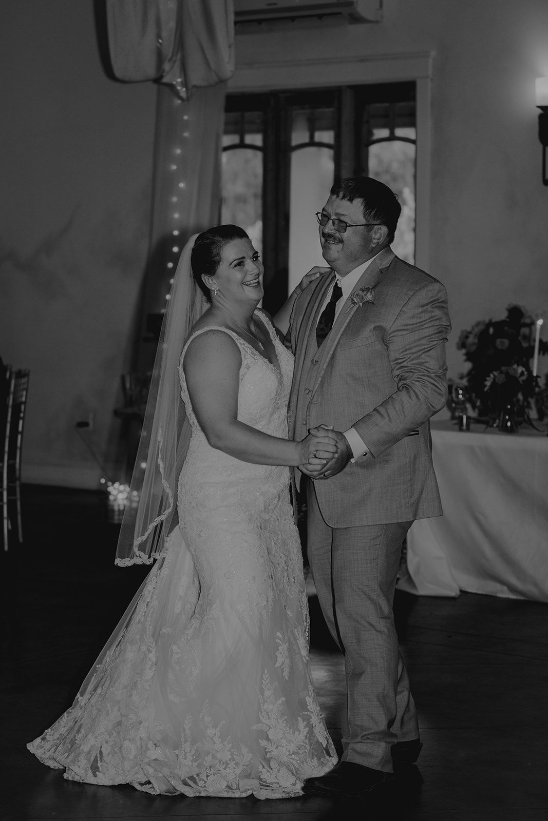 Bride in wedding dress dances with a man in a suit, both smiling, inside a venue.