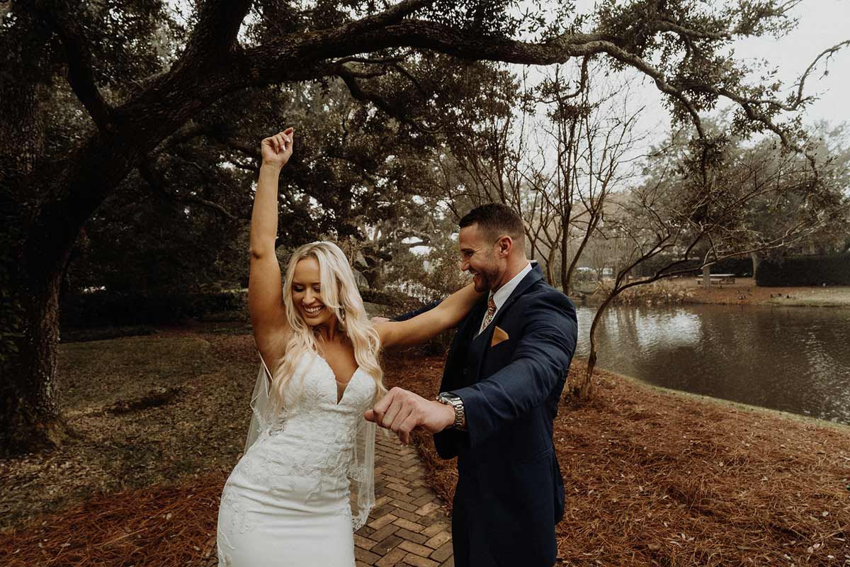 Bride in white dress raises arm, groom holds her hand; dancing outdoors near lake.