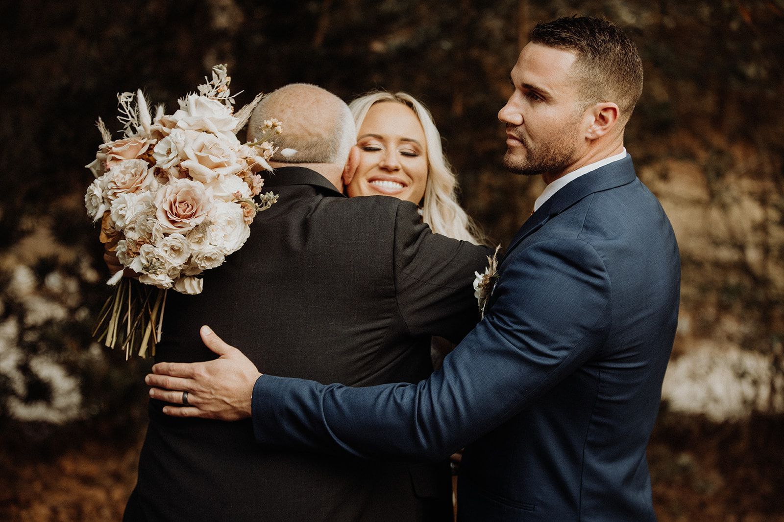 Bride hugs father with groom nearby; outdoor wedding, bouquet, happy smiles, forest background.