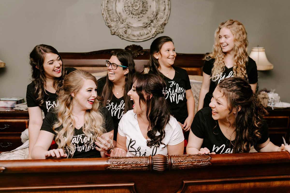 Bridesmaids in black shirts laughing together on a bed, with a floral wall hanging in the background.