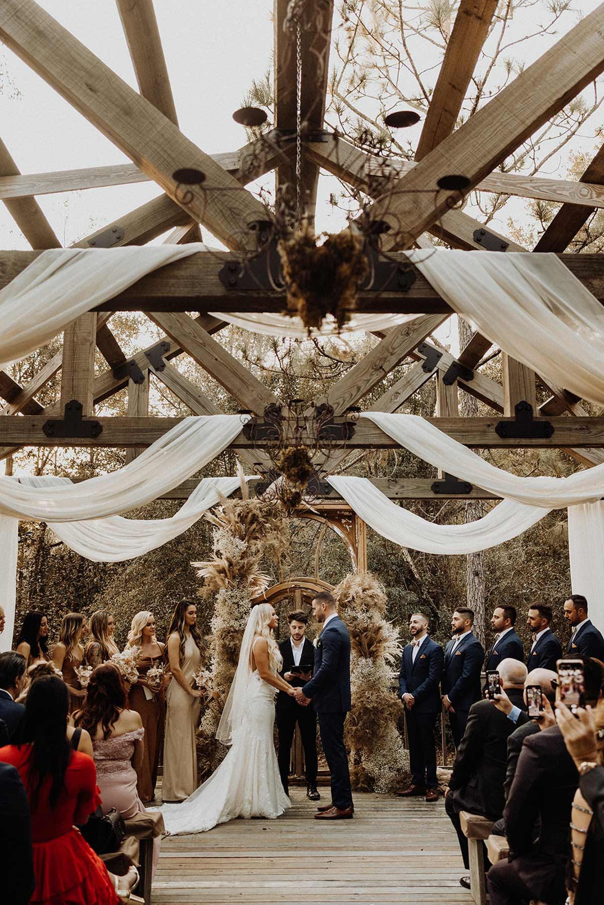 Wedding ceremony: couple at altar under wooden beams draped with fabric; guests watching.