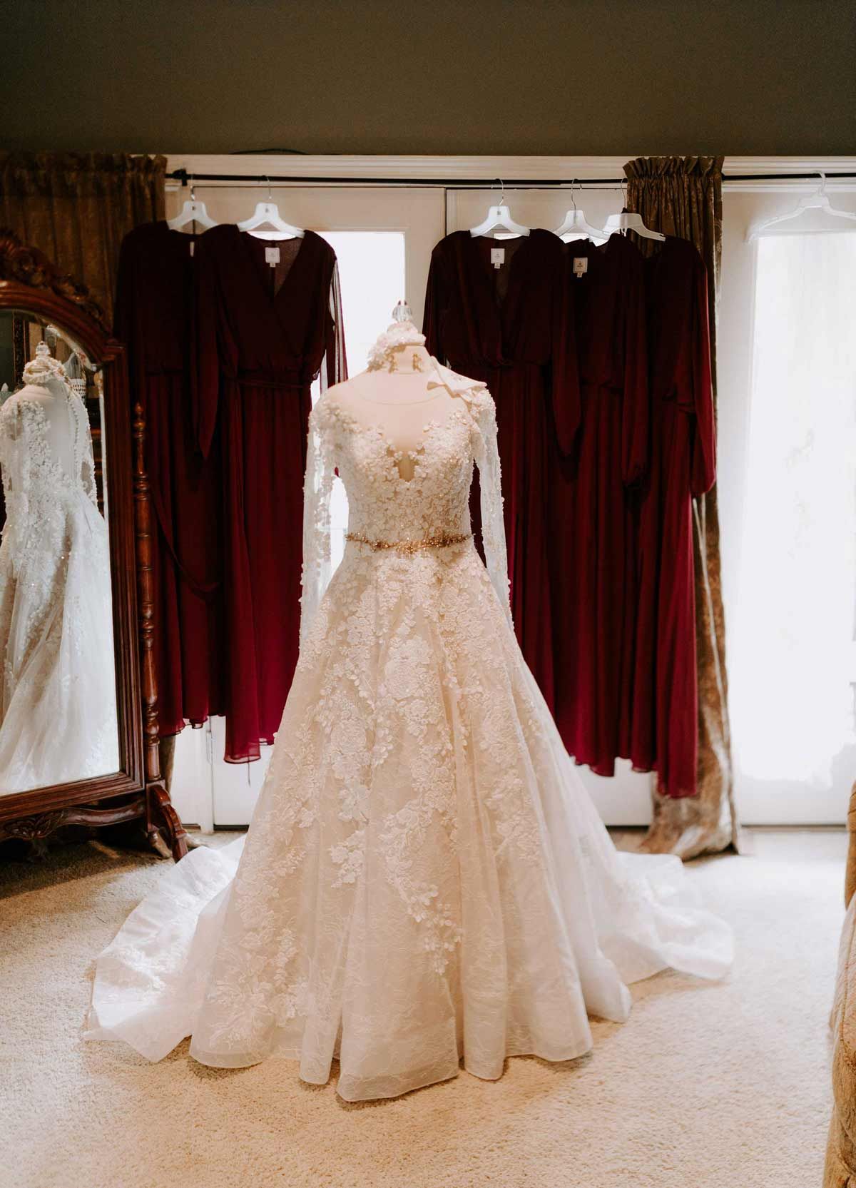 Wedding dress on a mannequin with burgundy bridesmaid dresses and a mirror in the background.
