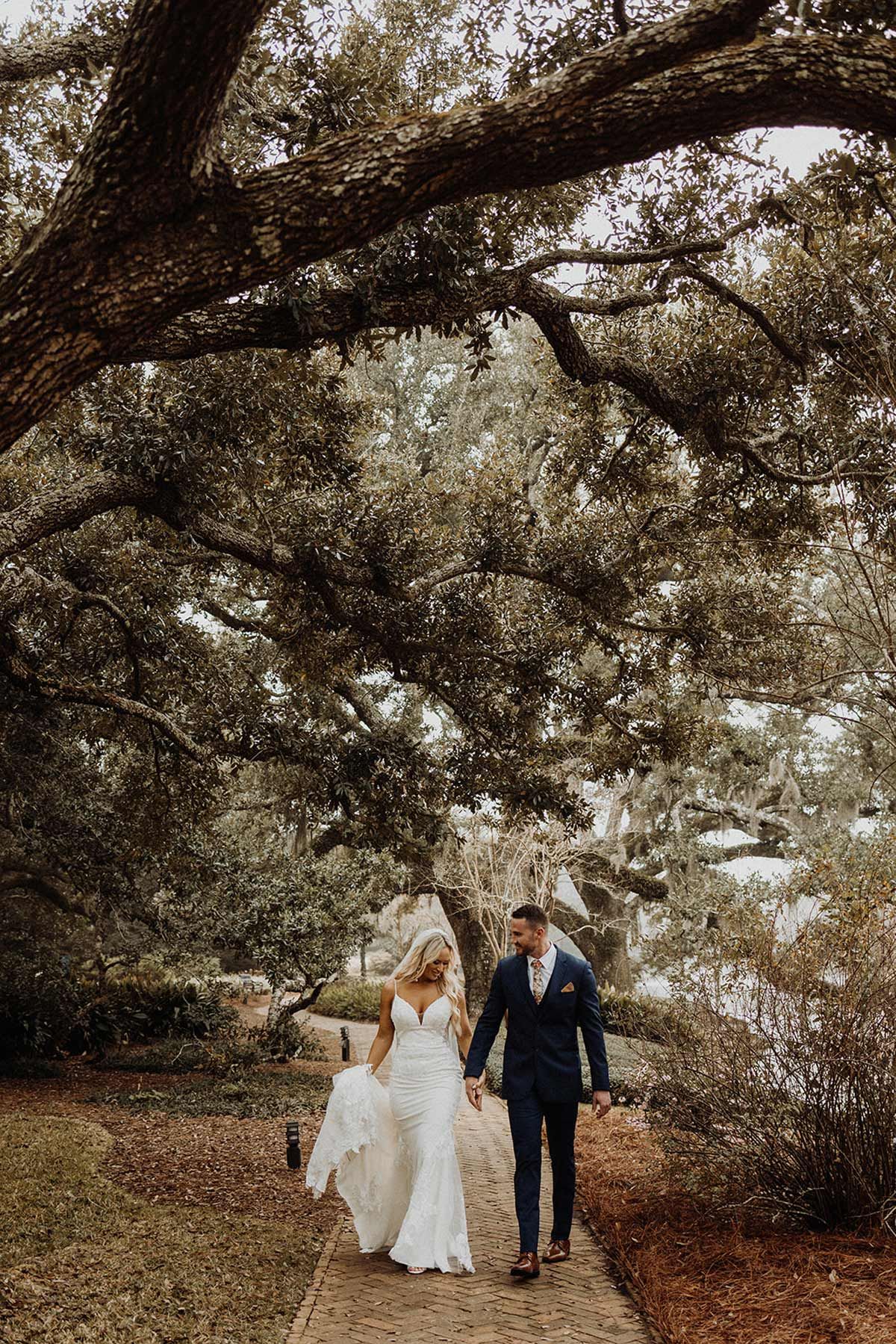 Bride and groom walking hand-in-hand on stone path under large tree; wedding day.