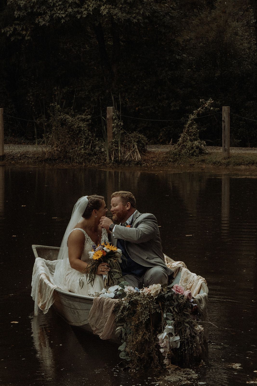 Couple in wedding attire on a rowboat in a pond, sharing a tender moment.
