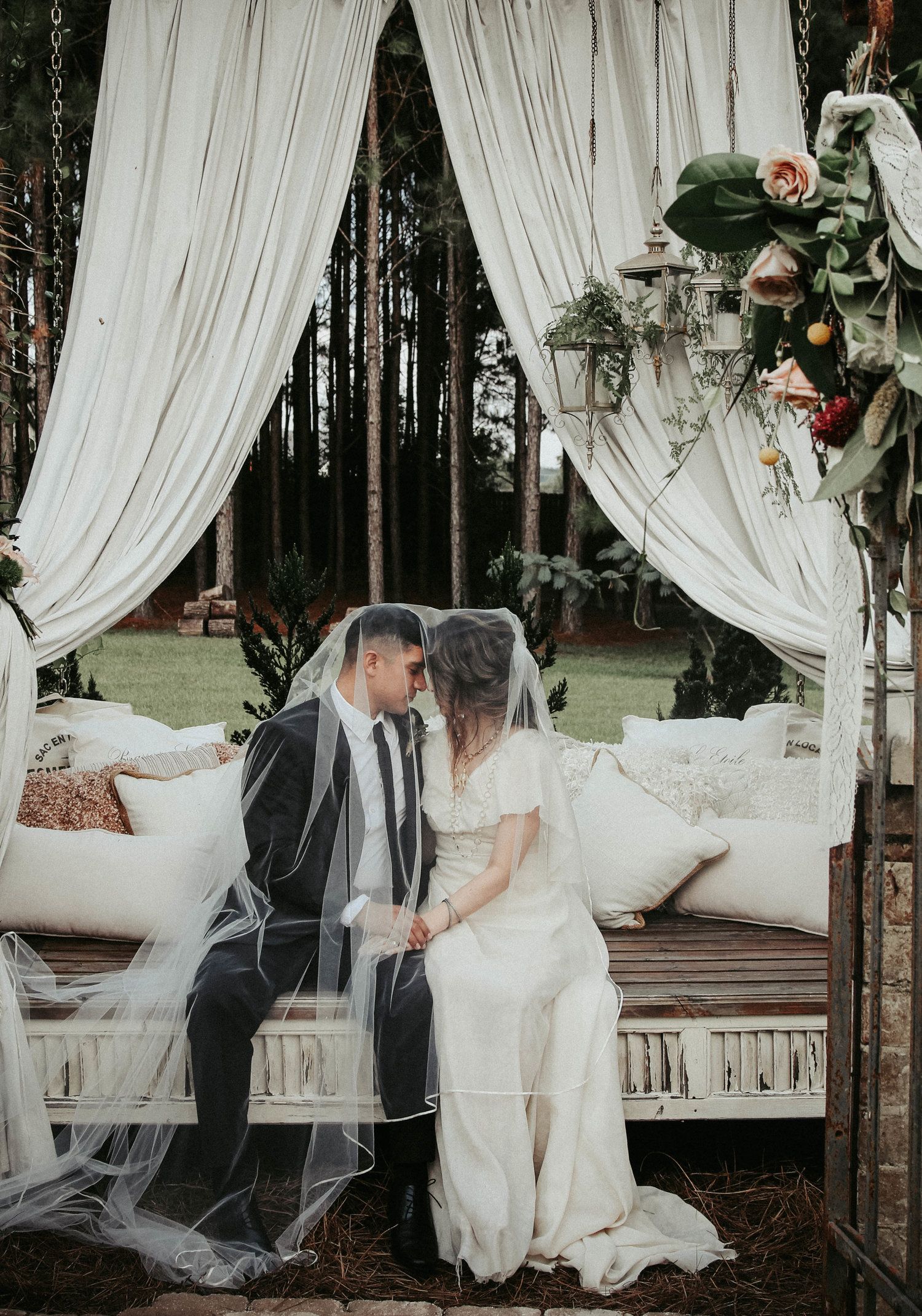 Couple kissing, sitting on a decorated bench draped with white fabric, in a wooded outdoor setting.