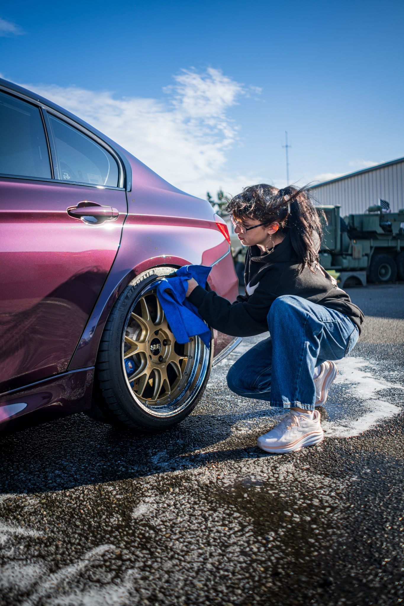 Person crouches by a maroon car, cleaning a gold wheel at a car wash under a blue sky.