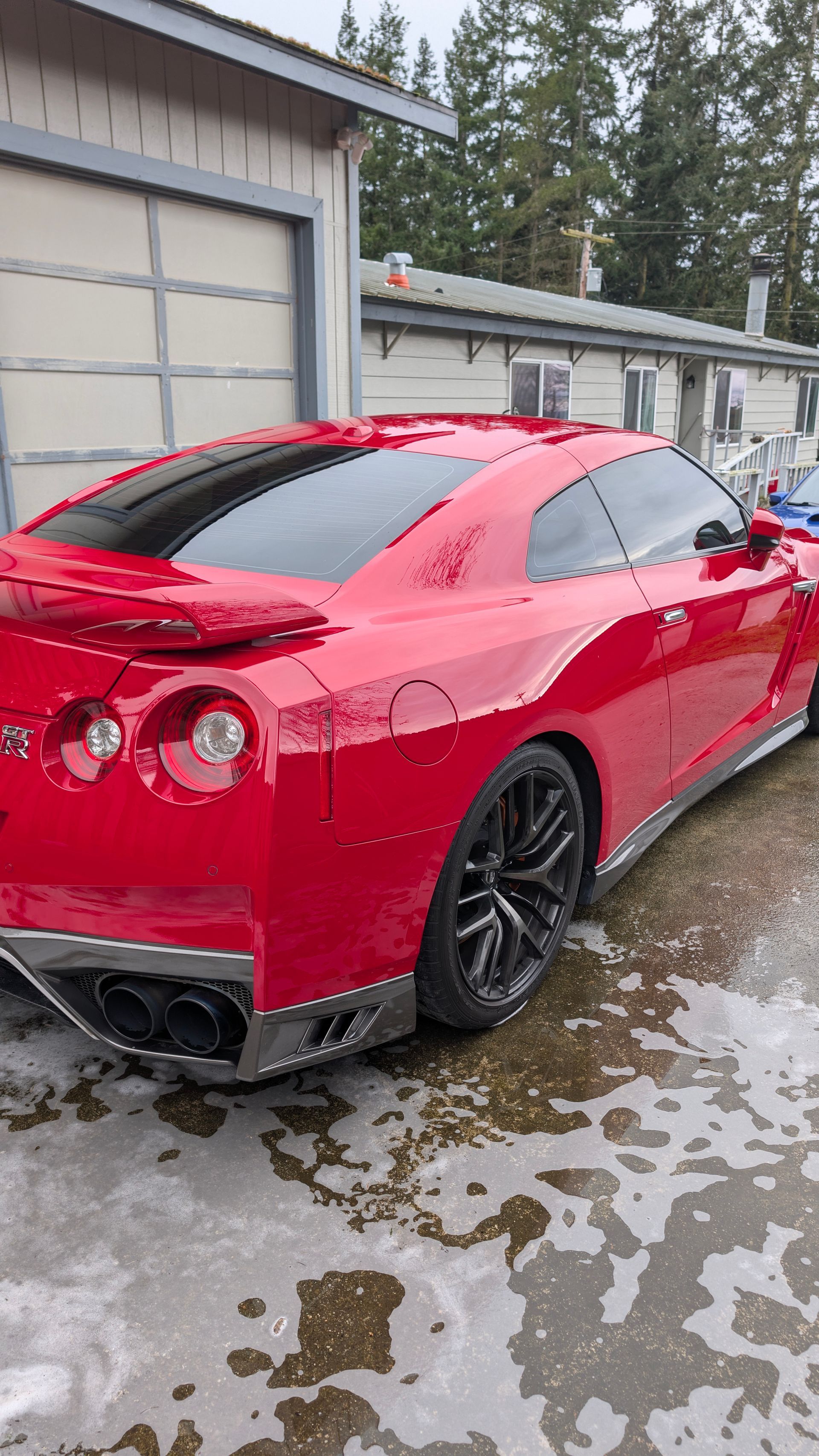 Red sports car parked on a wet driveway beside garage units, rear three-quarter view