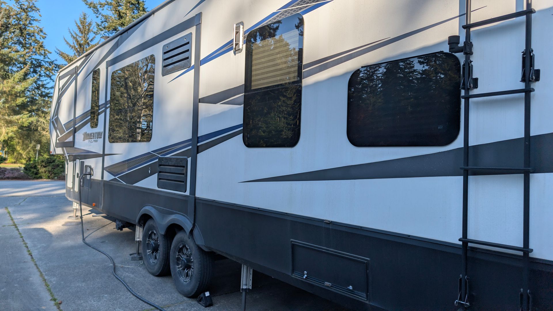 Large white and gray travel trailer parked on a sunlit driveway beside trees