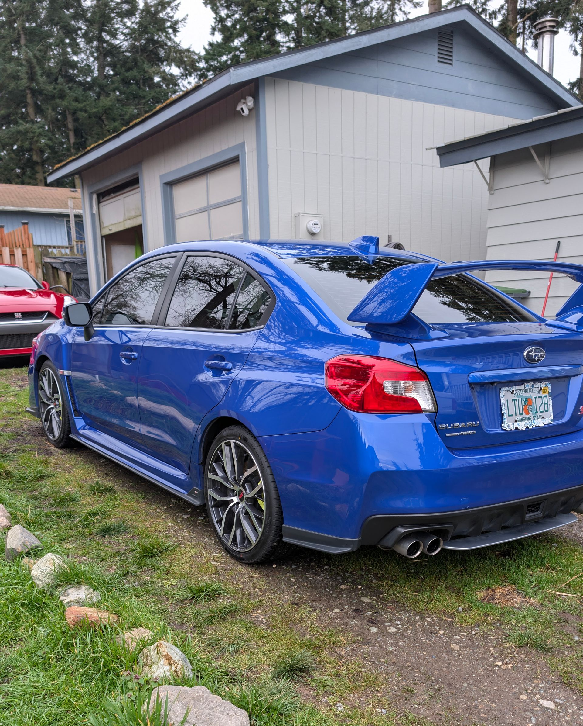 Blue sports sedan parked in a driveway beside a house, with a large rear spoiler.
